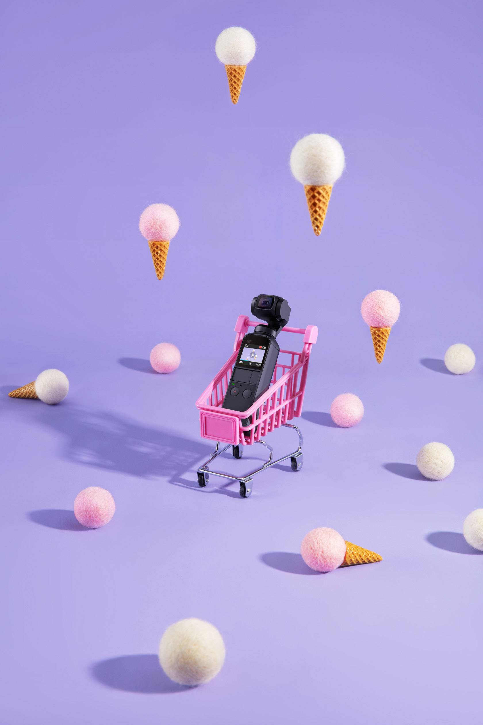 A small shopping trolley with a camera inside sits on a purple background, surrounded by floating ice cream cones and fluffy balls.