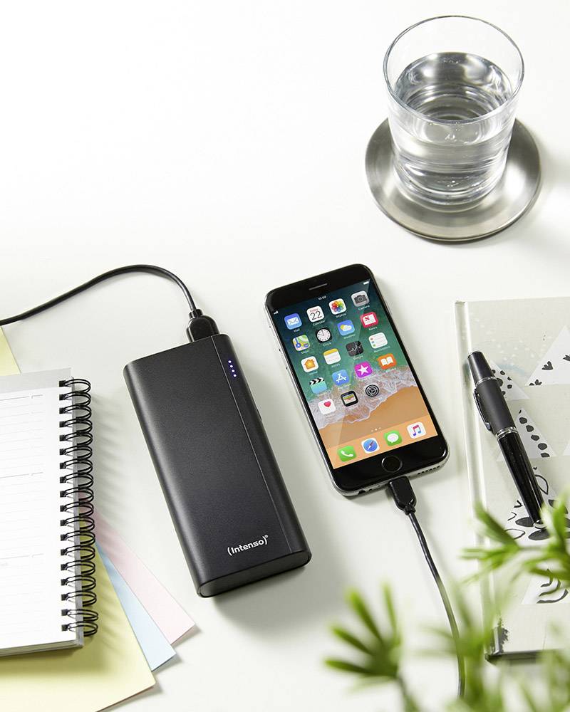 A smartphone is charging via a power bank. Next to it, there is a glass of water, a pen, and a notebook on a desk.