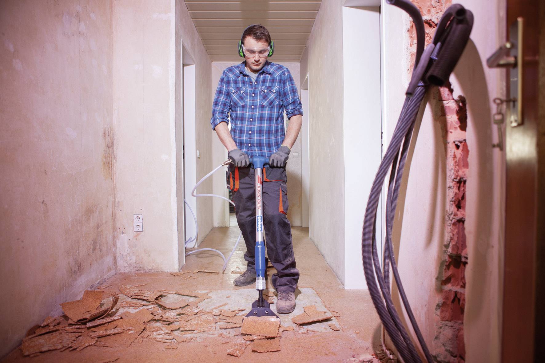 A man is working with a pneumatic hammer in a dilapidated hallway. Debris is scattered around him on the floor.
