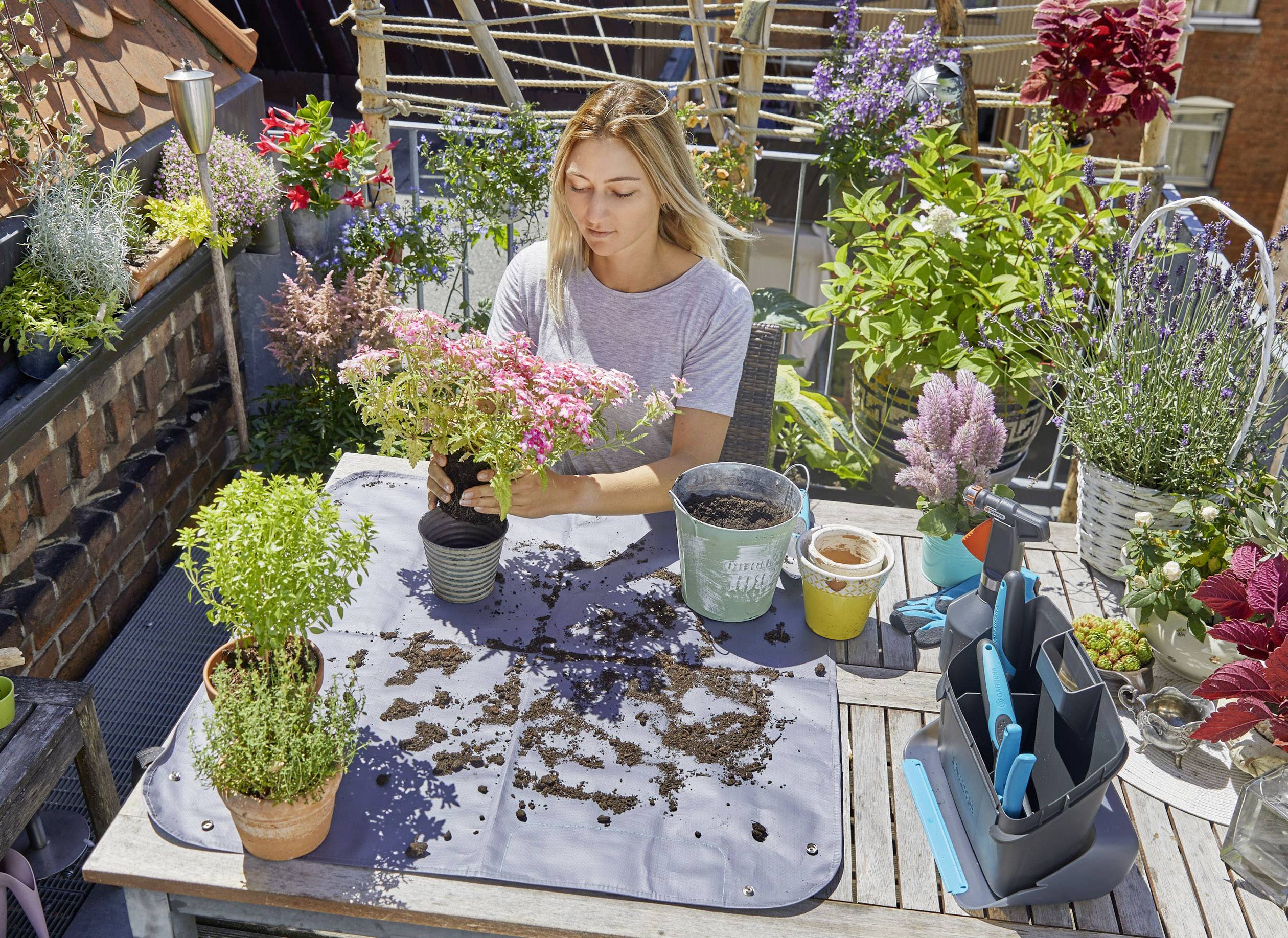 A woman is planting flowers on a balcony filled with green plants and colourful flowers in pots. She is preparing the soil on a table.