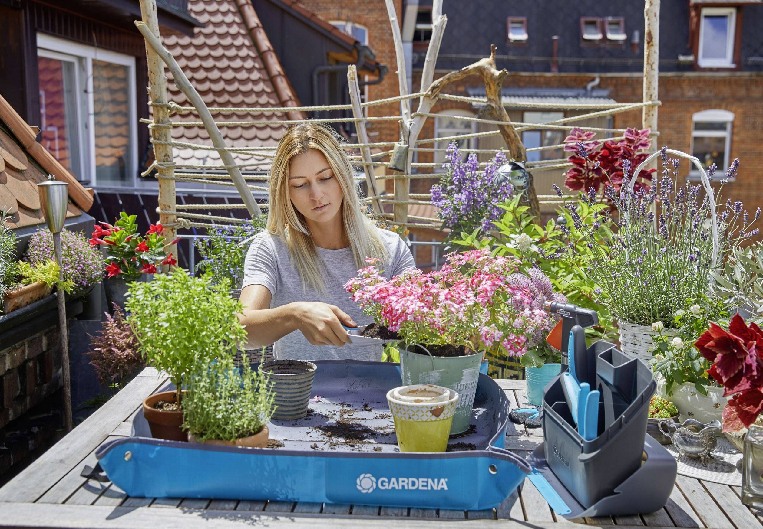 A woman is sitting at a table on a terrace, planting flowers in pots, surrounded by blooming plants and gardening tools.