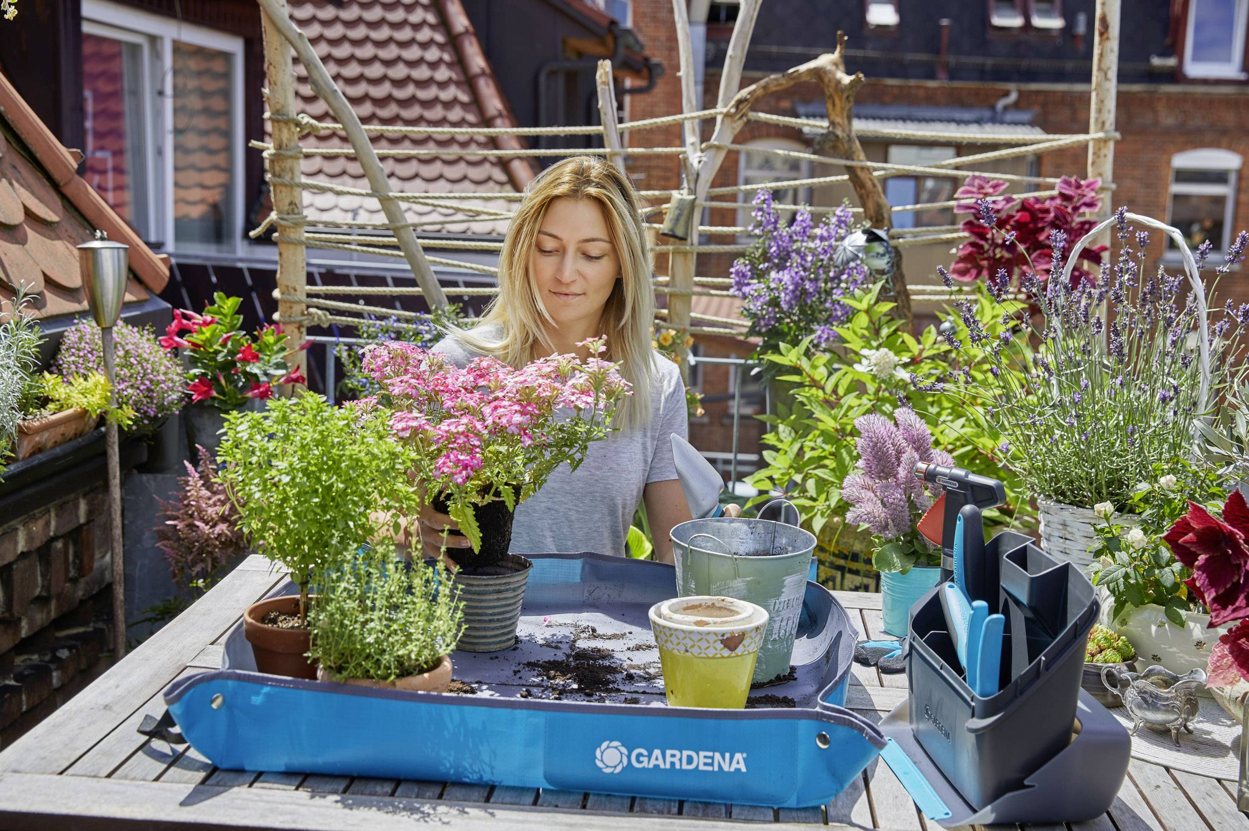 A woman is repotting plants on a balcony, surrounded by blooming flowers. A blue box of gardening tools sits on the table.