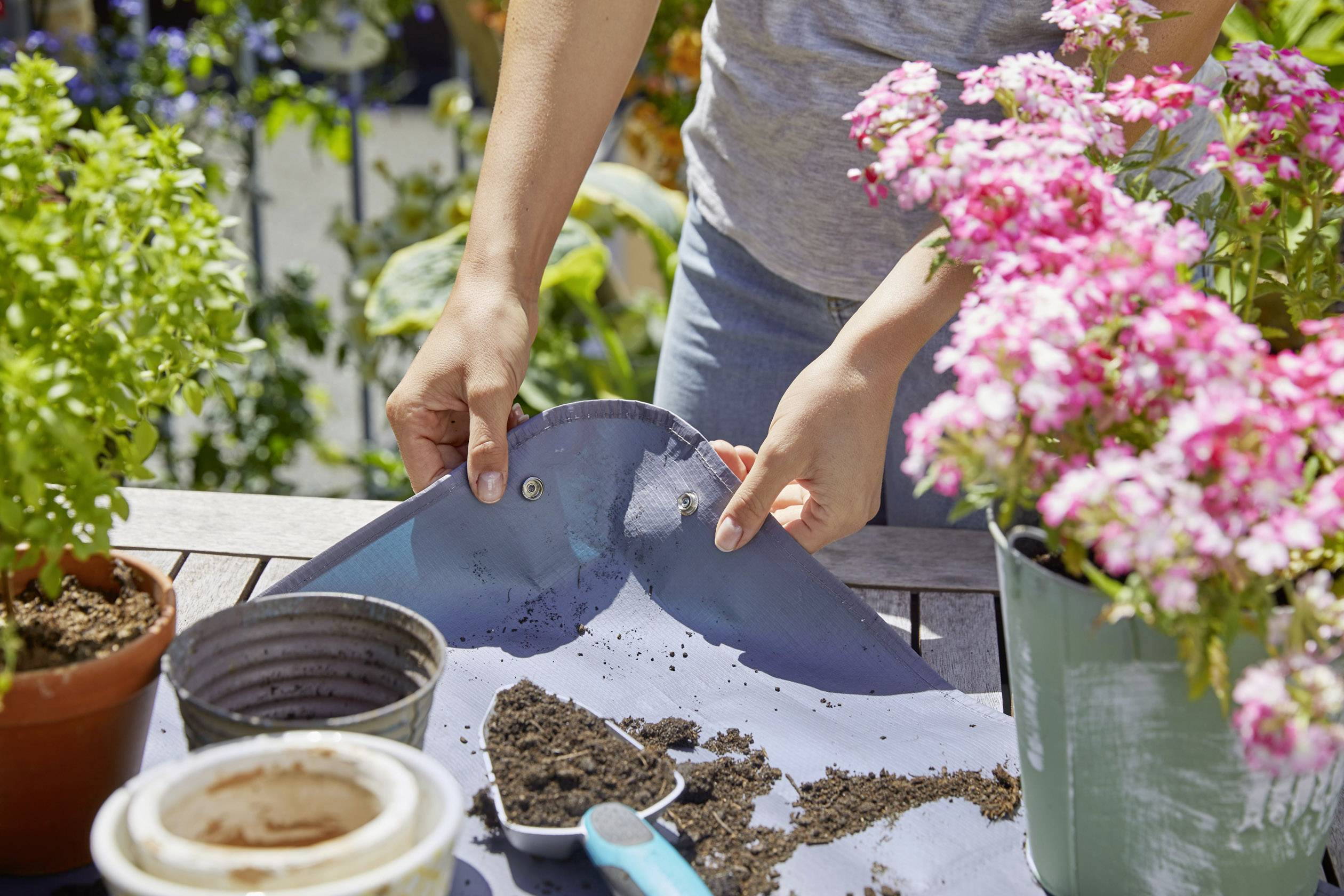 A person is filling soil into a pot on a table in a garden. Various plants are scattered around, including flowering pink blossoms.
