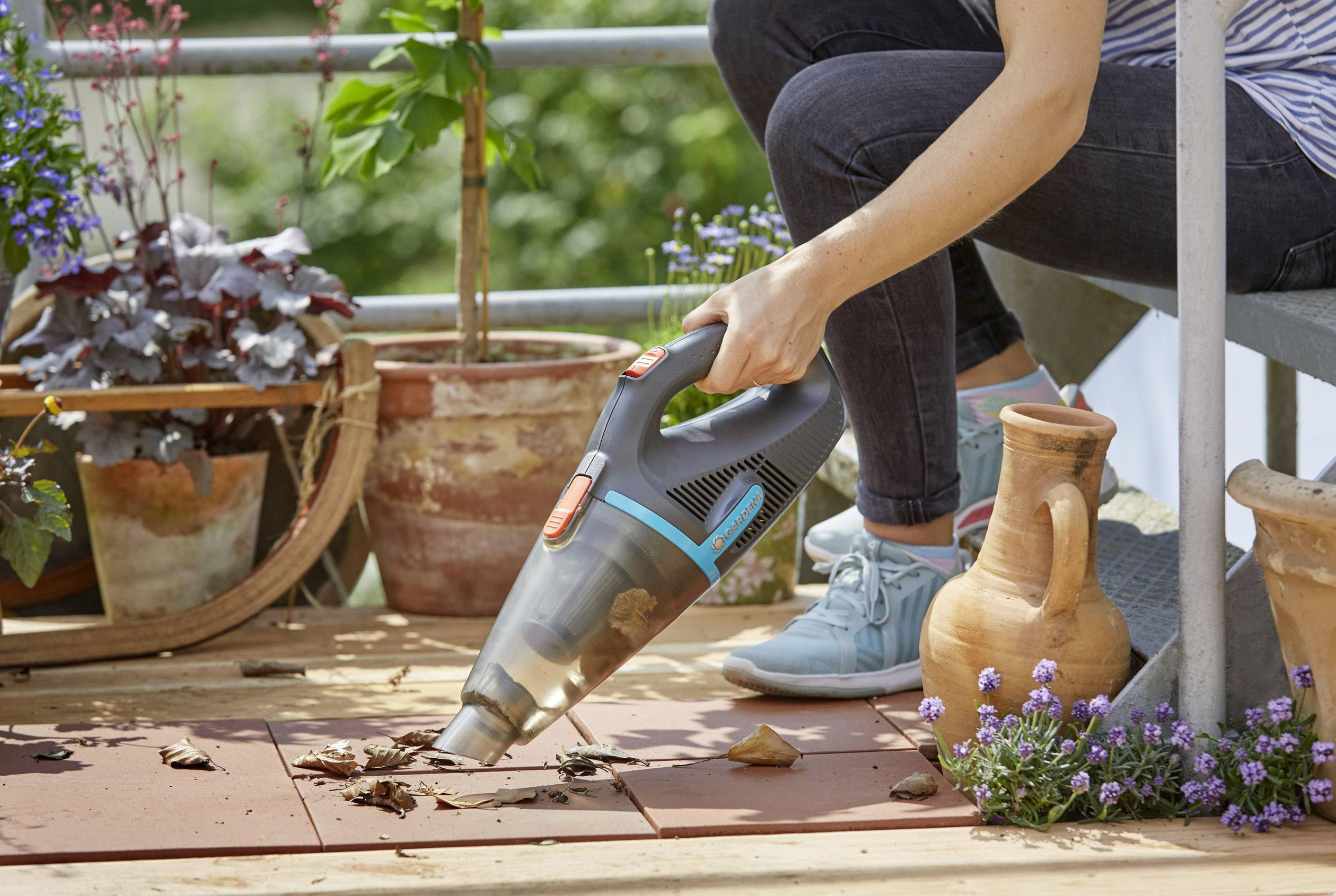A person is using a handheld vacuum cleaner to remove leaves and debris from a terrace with plants in the background.