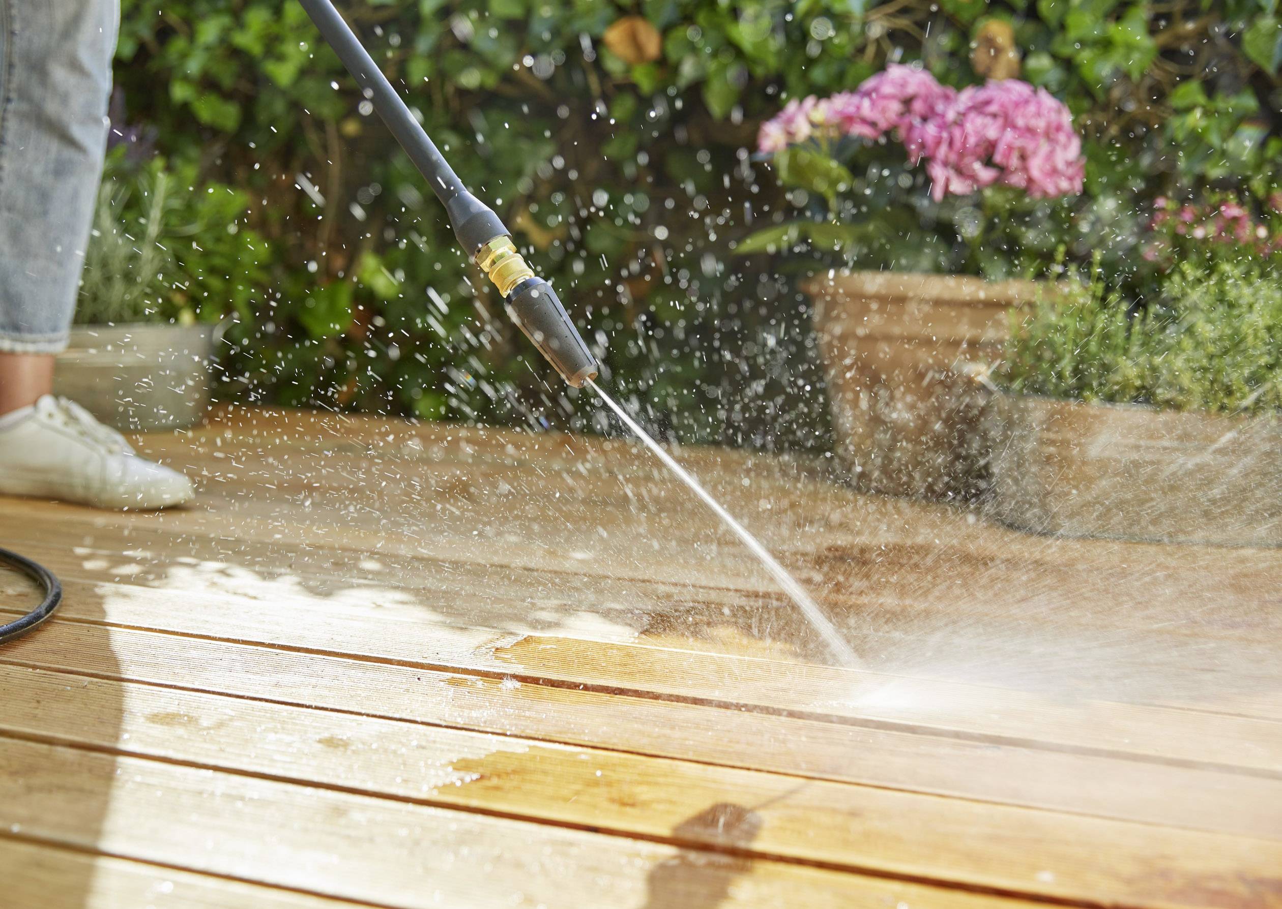 A person is cleaning a wooden decking with a pressure washer. Water is spraying in all directions. Plants are standing in the background.