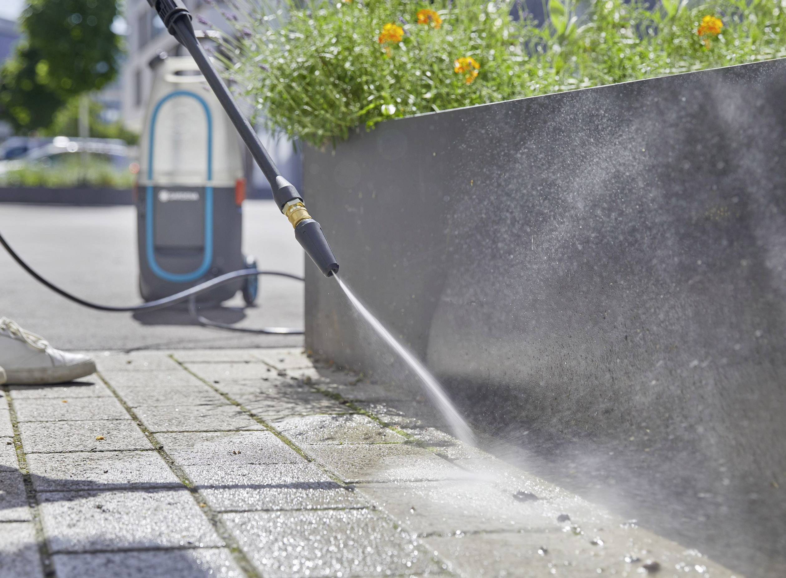 A person is cleaning paving stones with a pressure washer. Plants and a cleaning device are visible in the background.