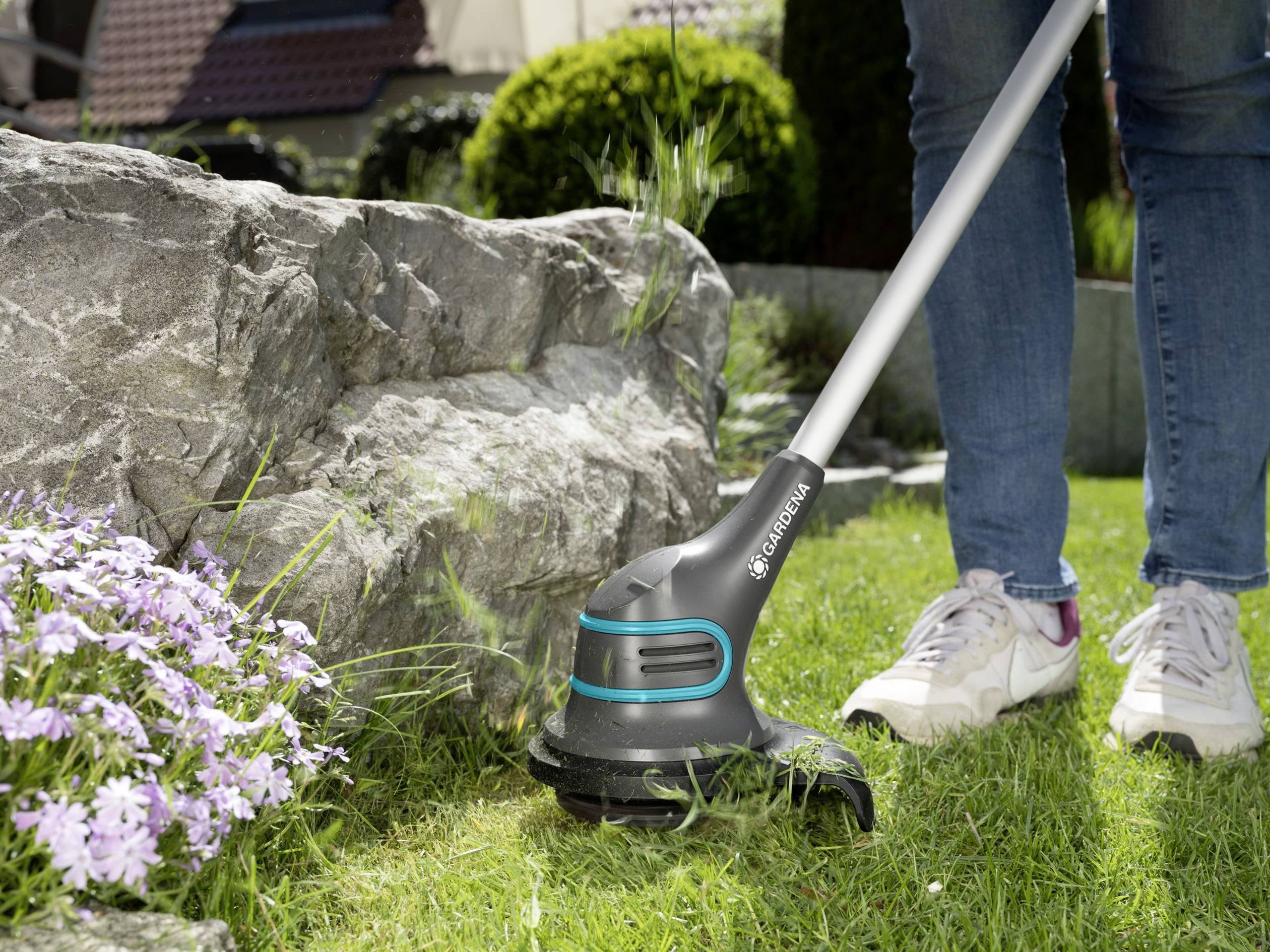 A person is trimming the edge of a garden with a cordless lawn trimmer, with purple flowers nearby. A large stone is positioned beside them.