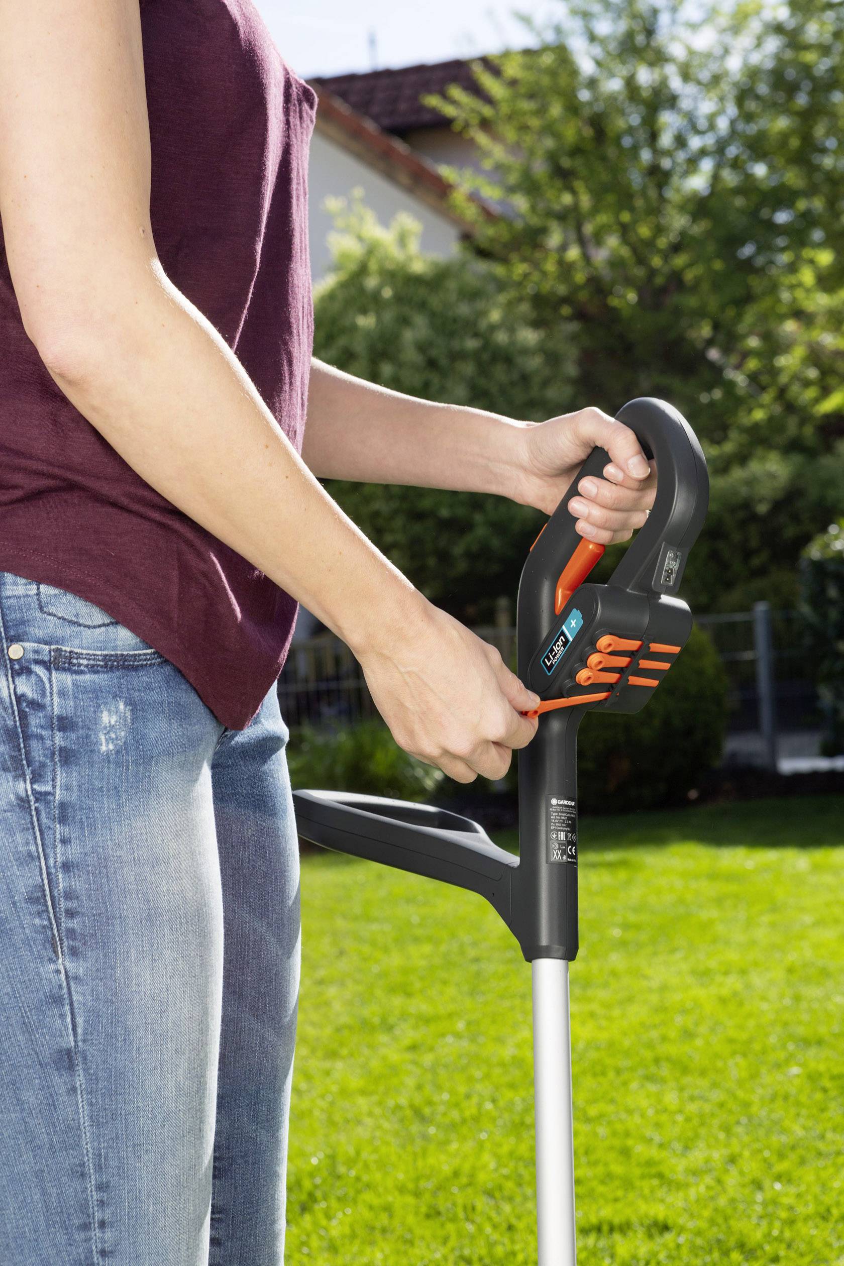 A person is using an electric garden trimmer on a lawn area. Trees and a fence are visible in the background.
