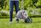 A person is standing on a lawn next to a garden hose reel. The background shows blooming plants.