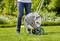 'Person standing next to a garden hose reel on a lawn, ready to water. Blurred plants can be seen in the background.'