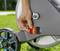 A hand operates the lever on a garden hose reel on a lawn. The wheel of the reel is partially visible.
