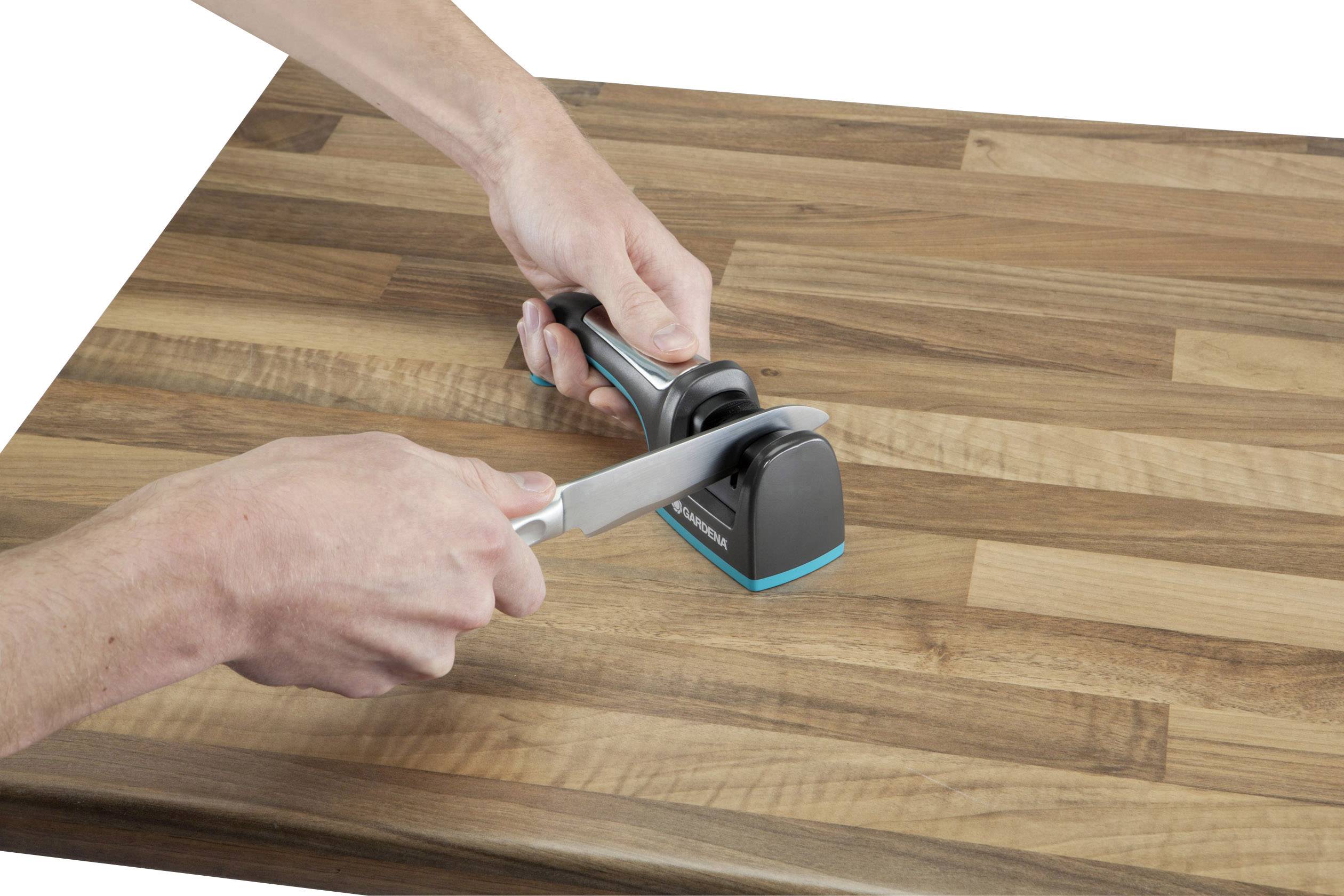 A person is sharpening a knife with a knife sharpener on a wooden board. Hands are holding both the knife and the sharpening device.