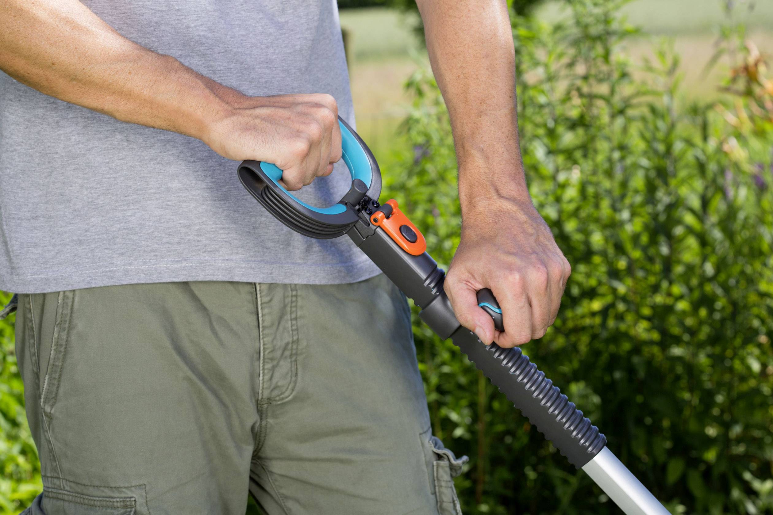 A person in a grey t-shirt and green trousers is holding the handle of a gardening tool outdoors, surrounded by plants.
