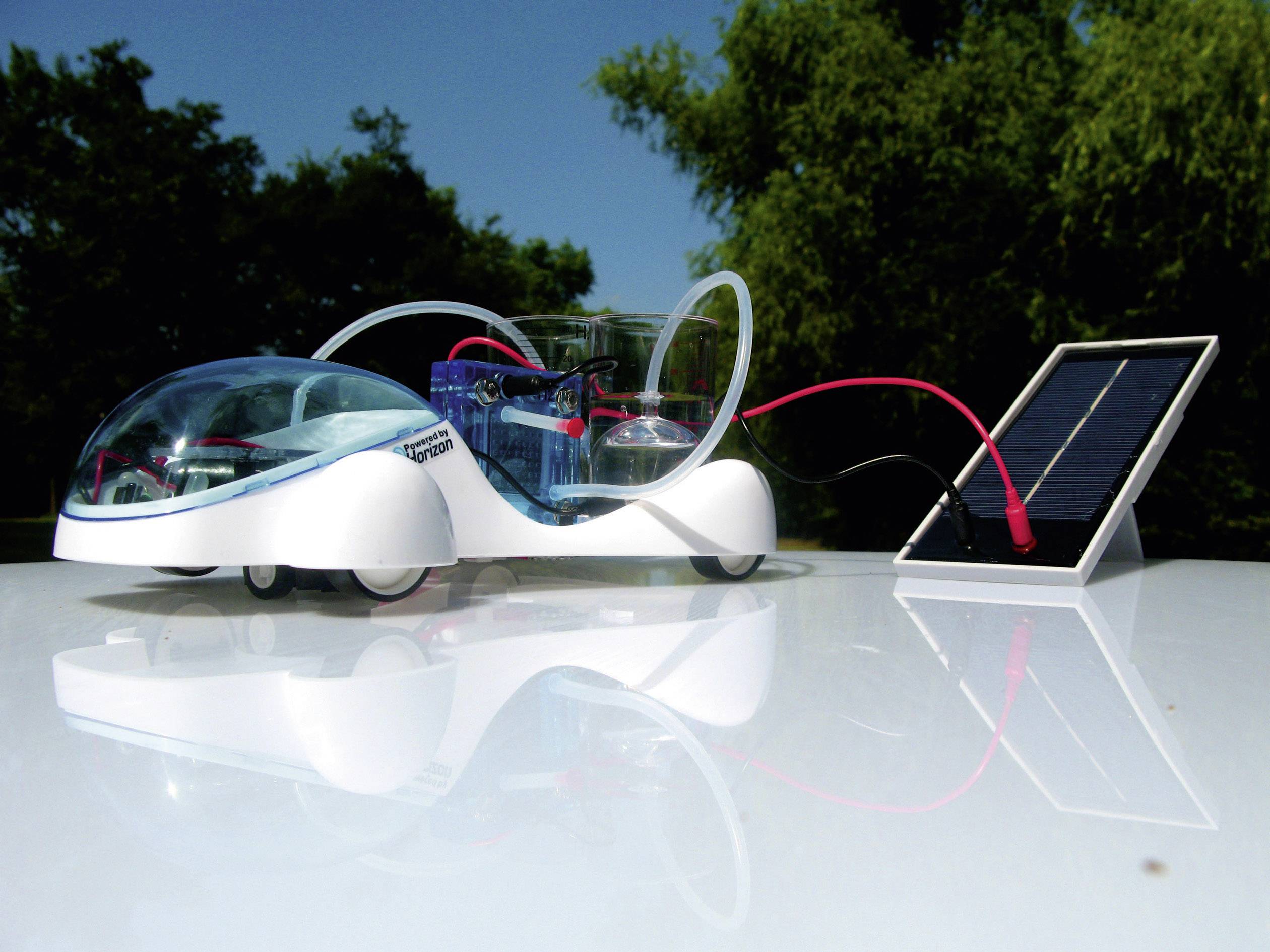 A small model car with a transparent cover, connected to a solar panel, stands under open skies.