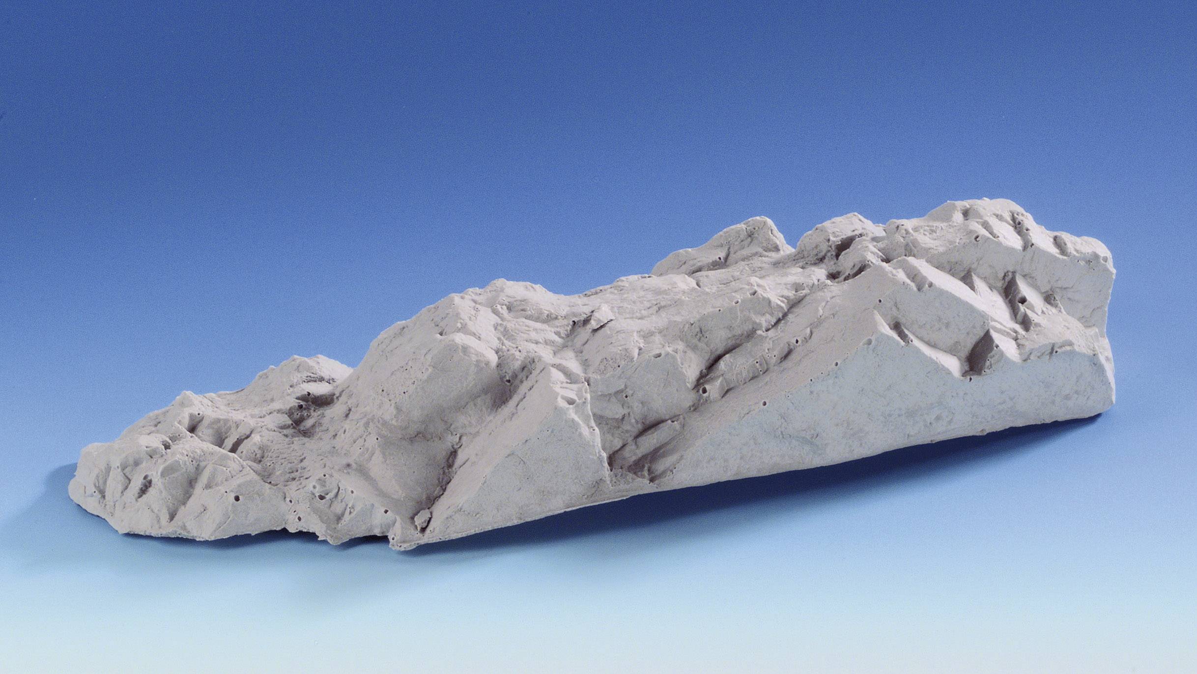 A grey, structural rock against a blue background. The rock comprises rough surfaces and edges that cast light and shadow.