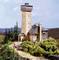 Model of a landscape with a tall tower and an adjacent building, surrounded by trees and small figures on a path.
