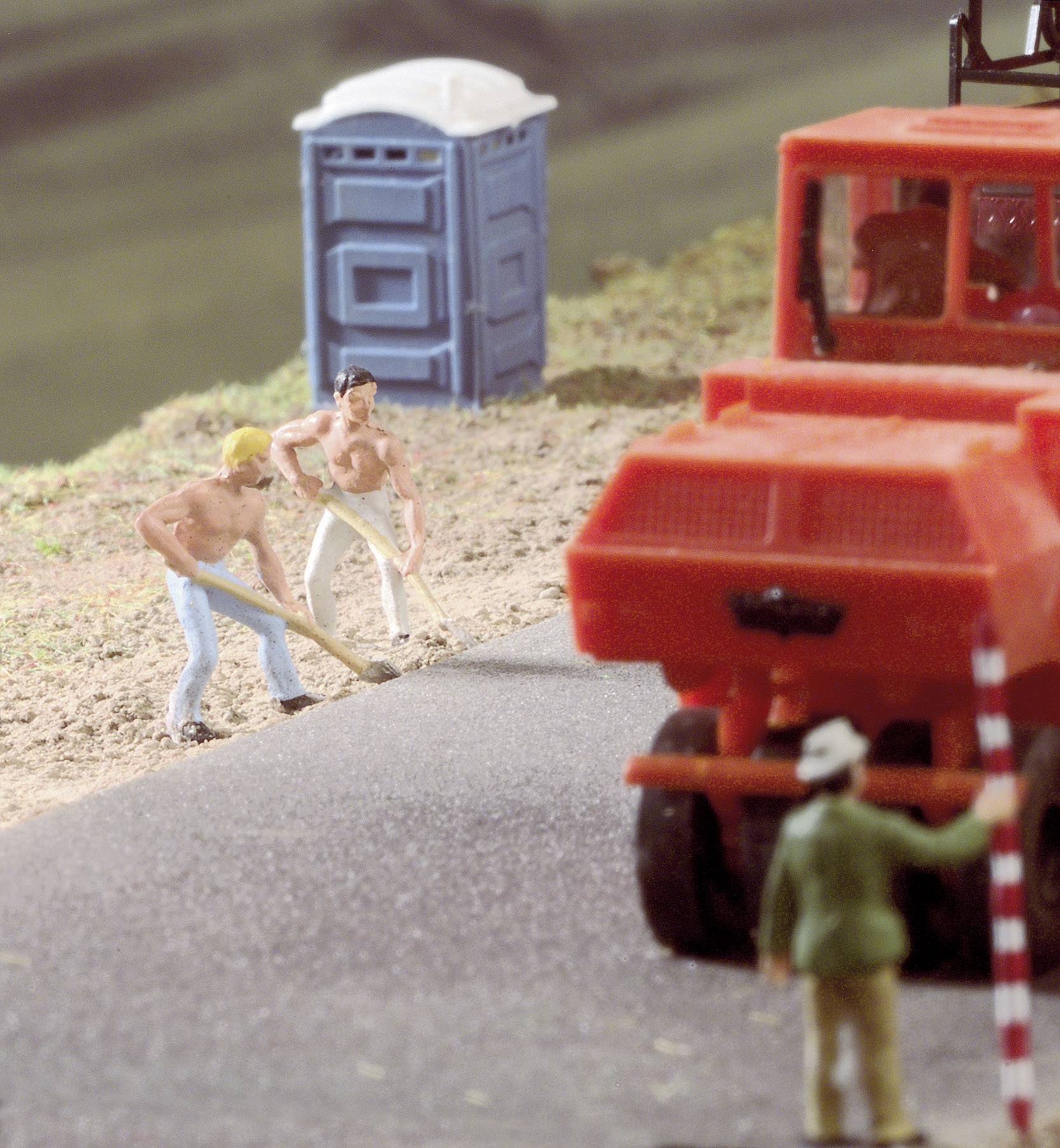 Miniature scene of construction work: Two workers in jeans digging earth beside a road, a red construction vehicle and a blue welfare unit in the background.