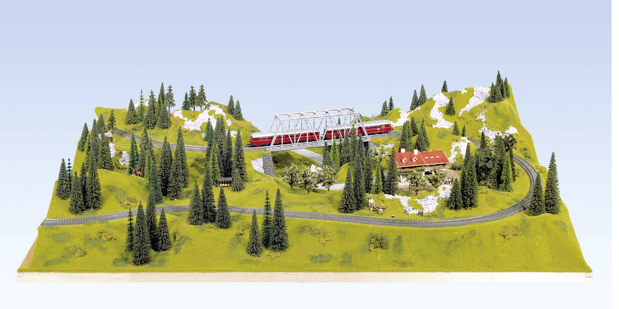 A model of a landscape with railway tracks and a red train on a bridge, surrounded by hills, forests, and small buildings.