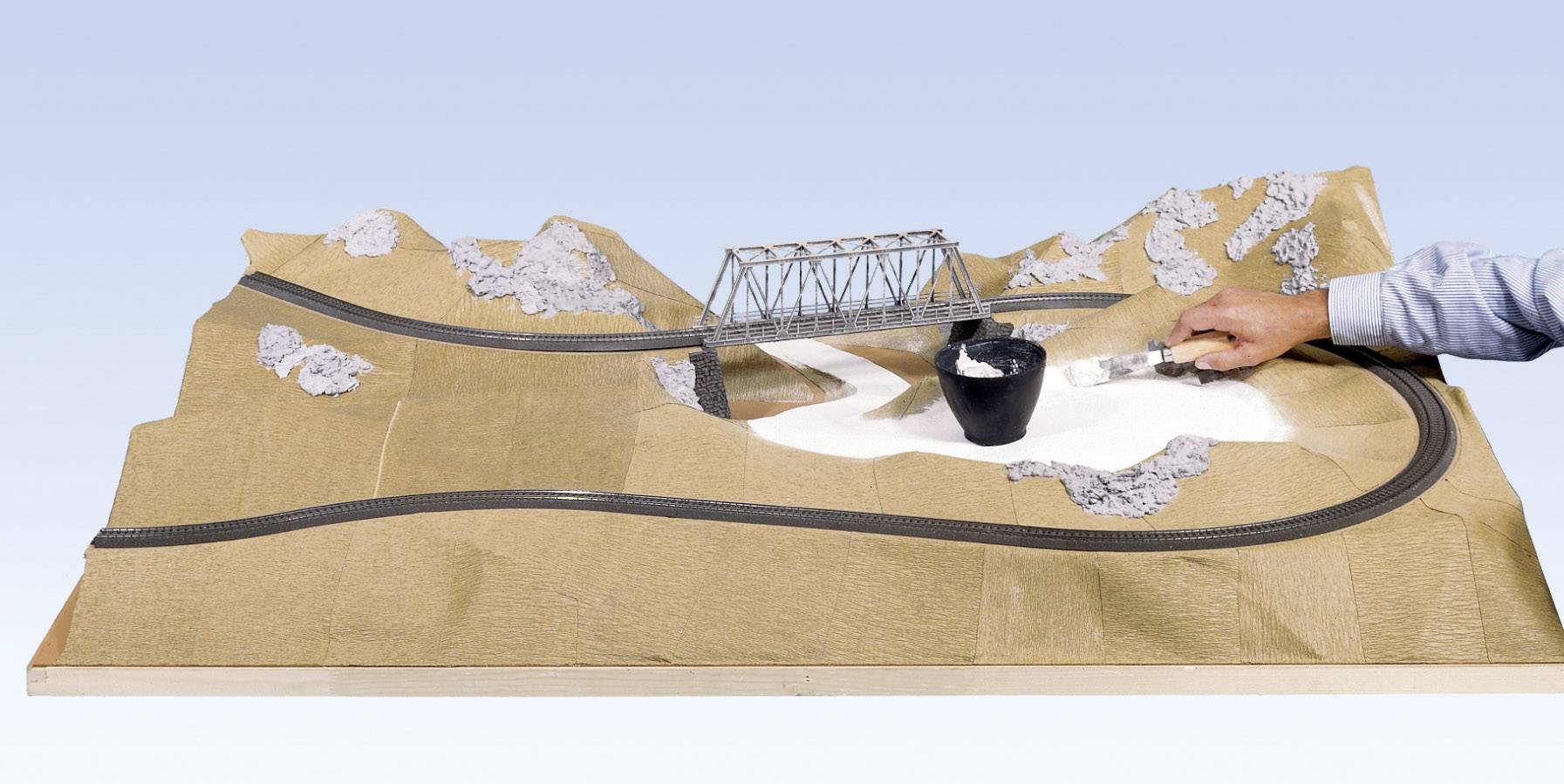 A person is painting plaster onto a landscape model featuring a bridge and railway tracks.