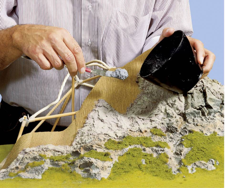 A person demonstrates the construction of a papier-mâché slope with a wire frame. Sand is being applied from a container to provide texture.