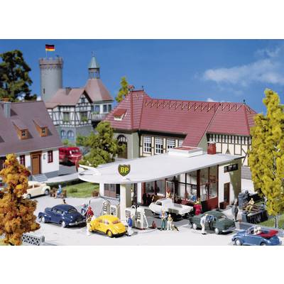 A miniature model town with a petrol station in the foreground, flanked by several vehicles and figures, near a castle.