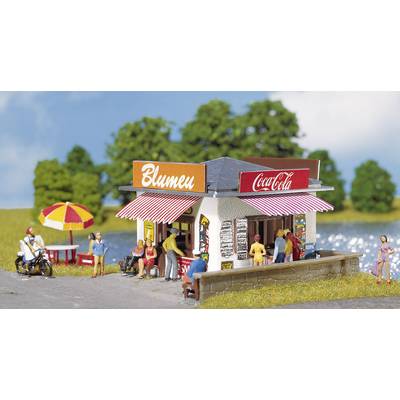 A small kiosk by the lakeside with the signs 'Flowers' and 'Coca-Cola', surrounded by people and trees under a blue sky.