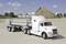 A white lorry with a trailer, loaded with large pipes, is parked on a gravel area. An earth mound is visible in the background.
