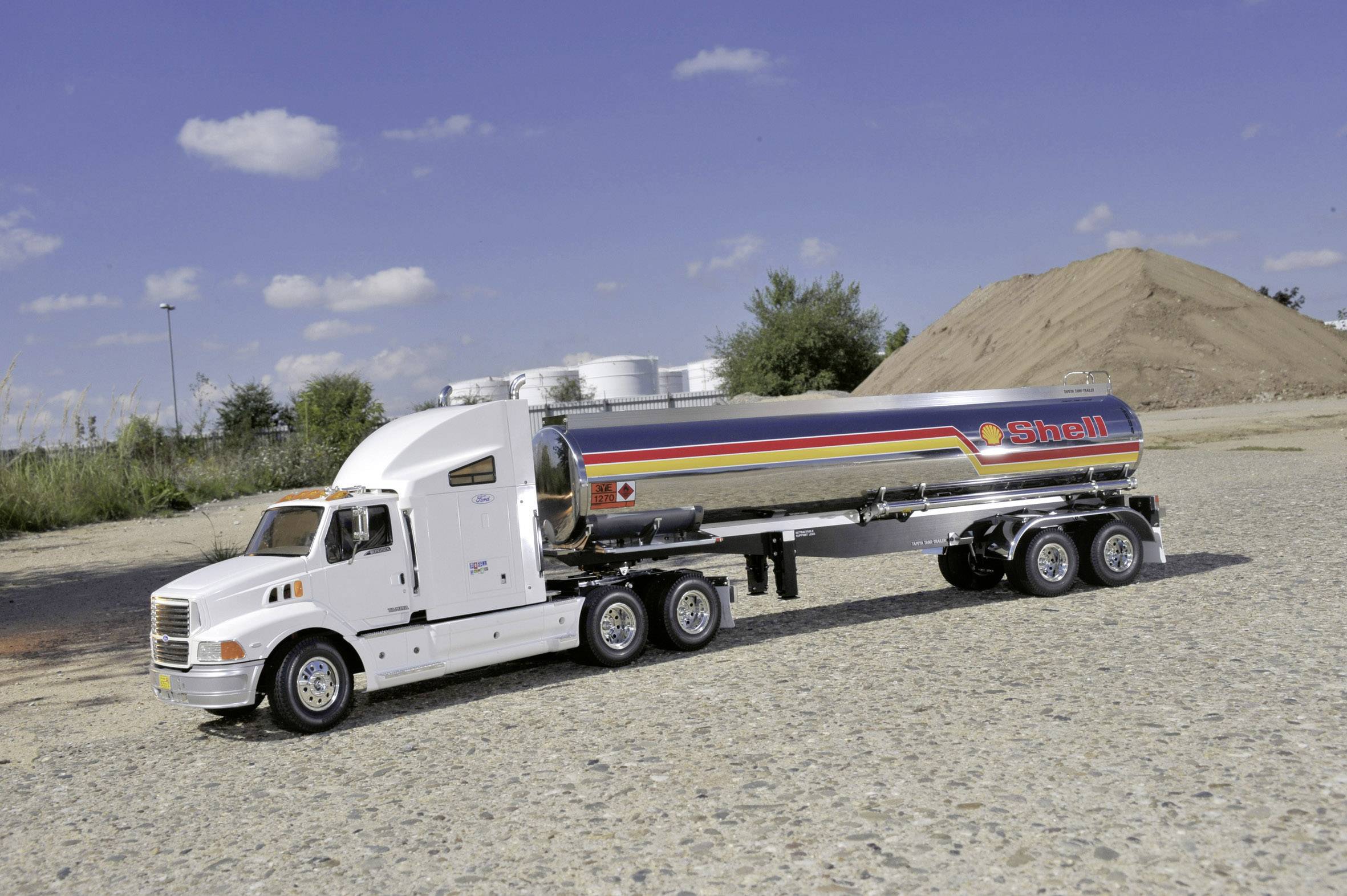 A Shell fuel tanker is parked on a construction site in sunny weather, surrounded by an earth mound and trees.