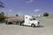 A white fuel tanker is parked on a gravel road beneath a blue sky. Trees and buildings are visible in the background.