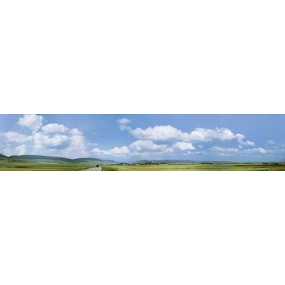 Panorama of a green landscape with expansive fields beneath a blue sky dotted with white clouds. Hills are visible on the horizon.