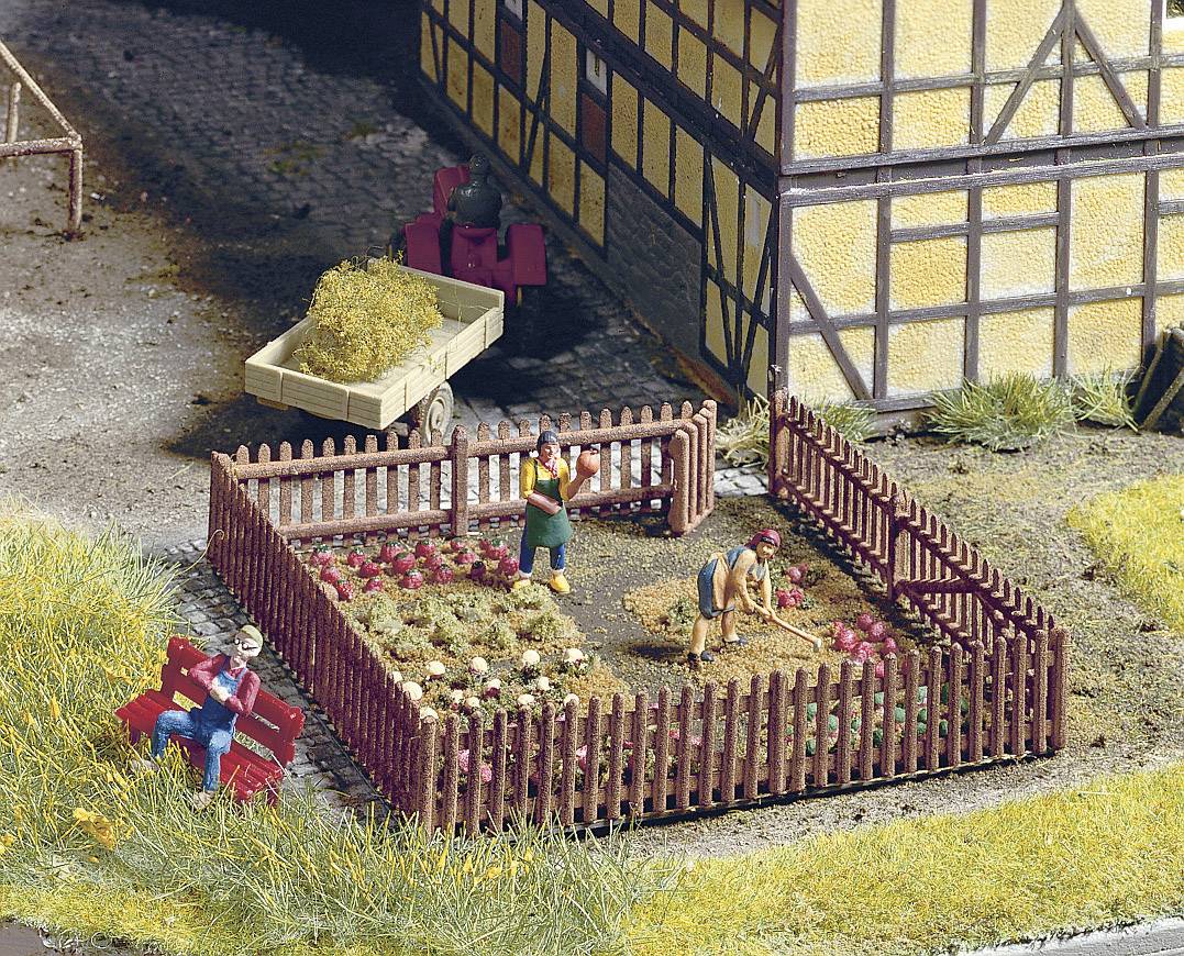 A small cottage garden with a fence, where two people are planting vegetables. In the background, a trailer with hay bales stands.