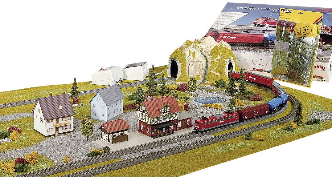 A model train travels through a miniature landscape with houses, trees, and a tunnel. In the background, a package of accessories.