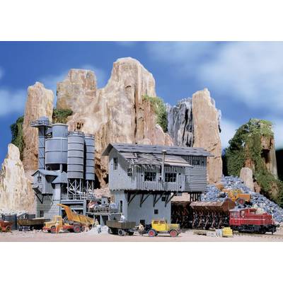 Model railway scene with an industrial facility set against a rocky landscape. Main building with silos, surrounded by miniature vehicles and a train.