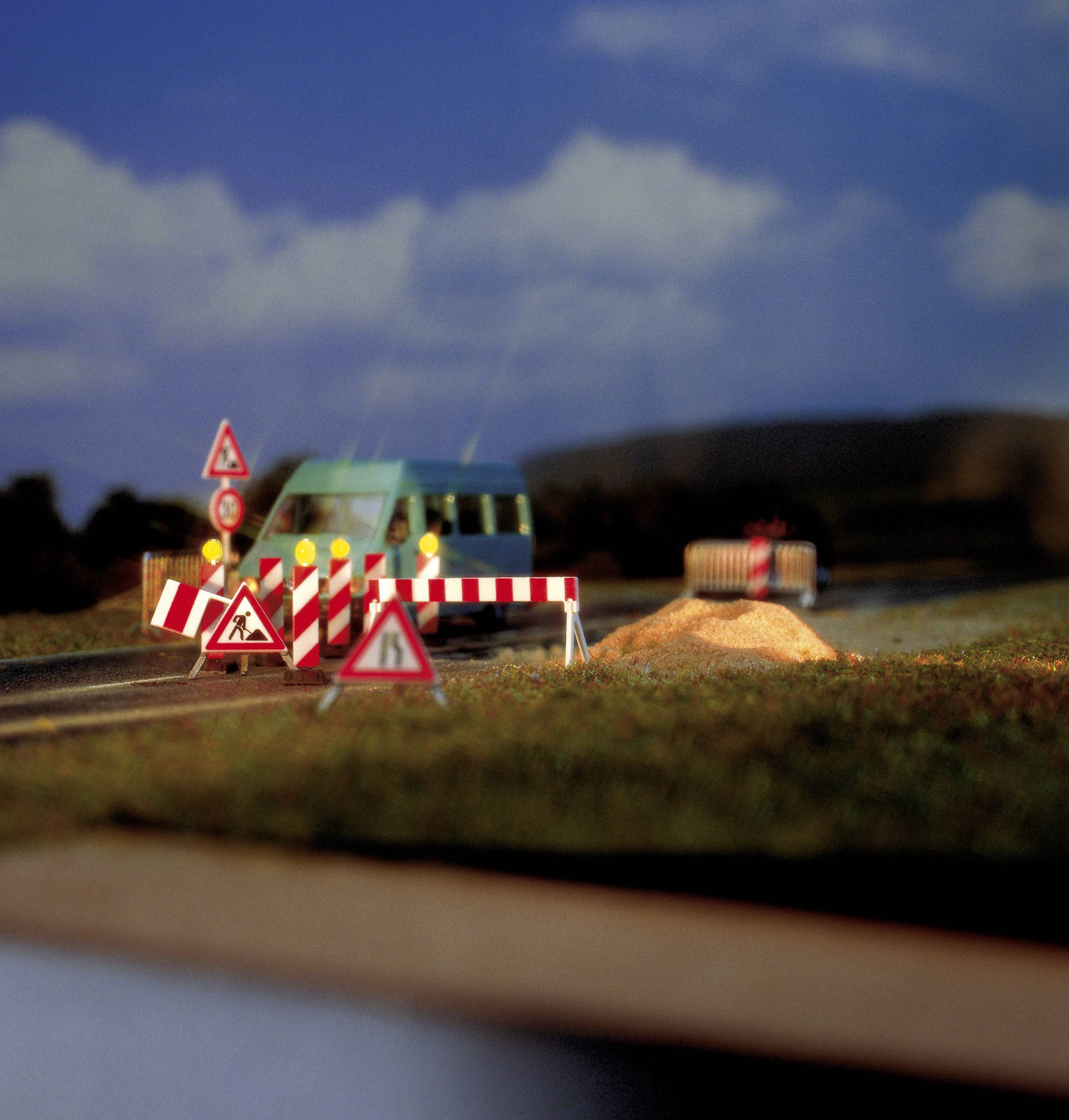 A model car is positioned in front of a roadblock with traffic signs and sand piles on a country road. The background shows a cloudy sky.
