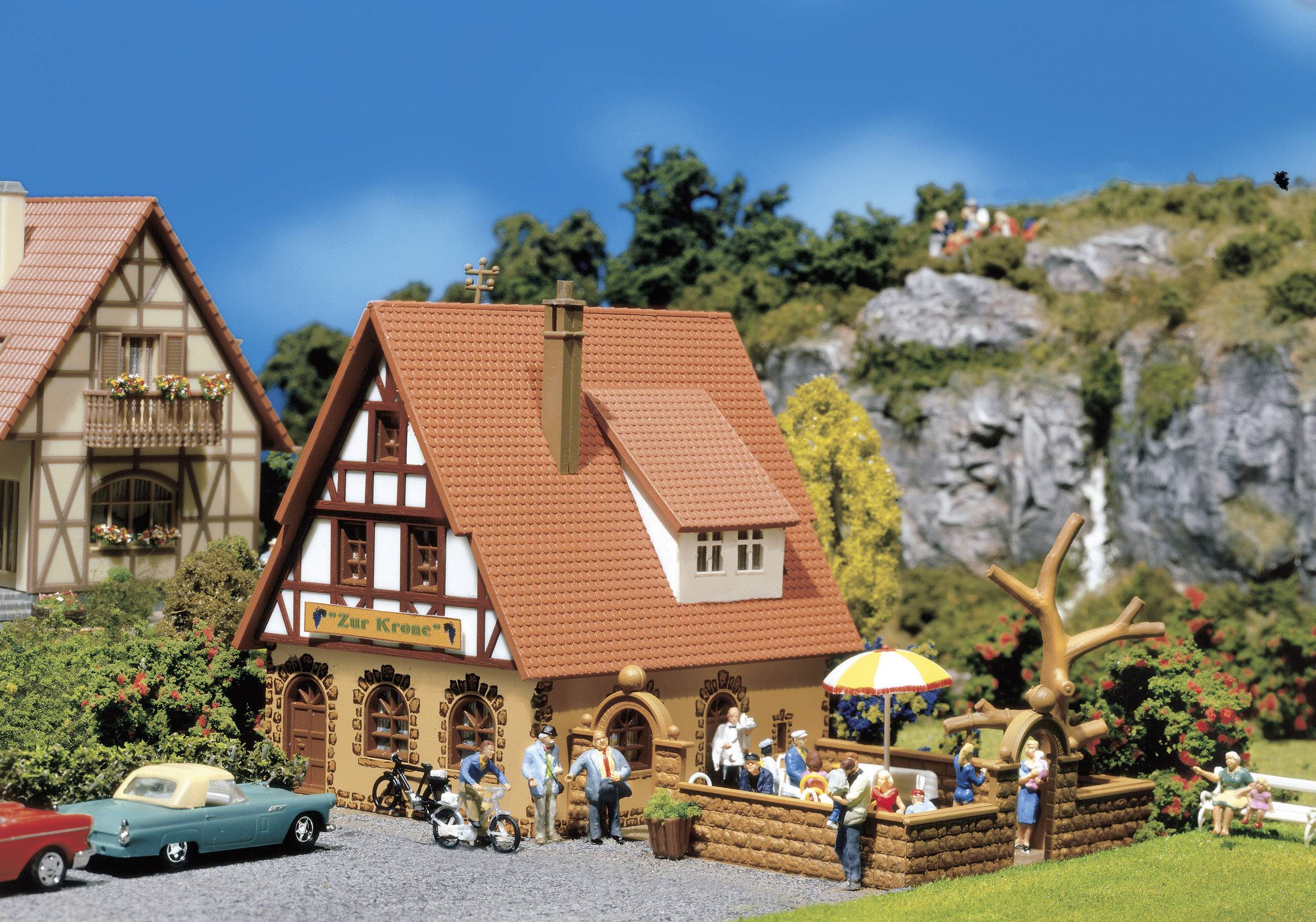 A miniature village depicts half-timbered houses with red roofs, people having a picnic and bicycles. Hills and a waterfall are visible in the background.