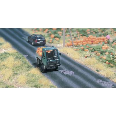 A small model lorry is transporting pumpkins along a country road, followed by a model car. The surrounding environment features fields and pumpkins.