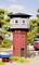 A red, round water tower with a grey upper storey stands in a rural setting with trees and a passing train.