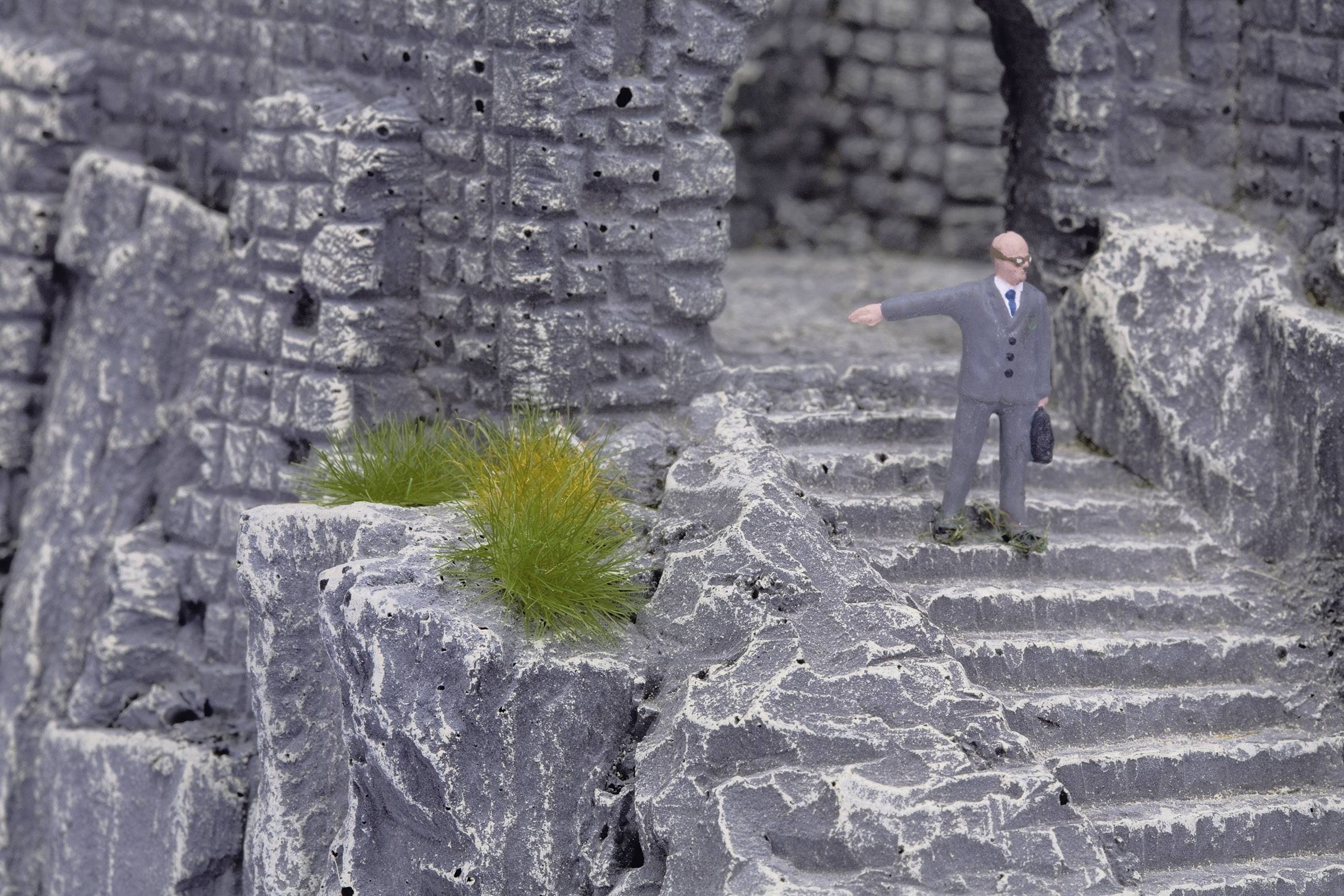 A miniature figurine of a man in a suit stands on a stone staircase of an old, ruins-like structure.