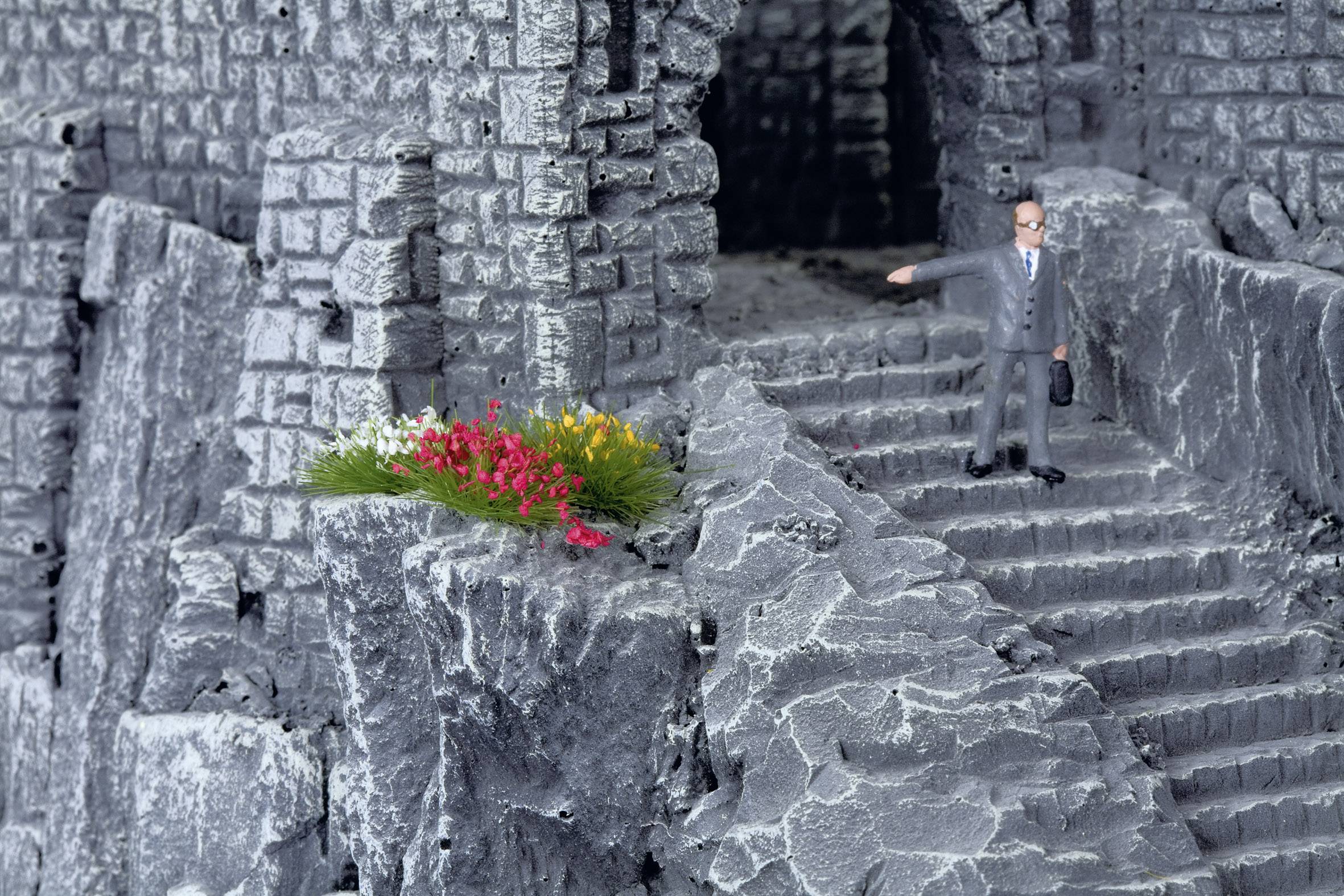 A miniature figure of a suited individual stands on grey, stone steps in a rocky, ruin-like environment. To the left, a small bouquet of flowers blooms.