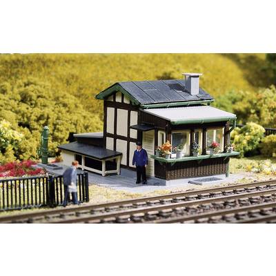 Small model railway house with half-timbered design and flowers on the windowsill, two figures beside it, in front of a level crossing.