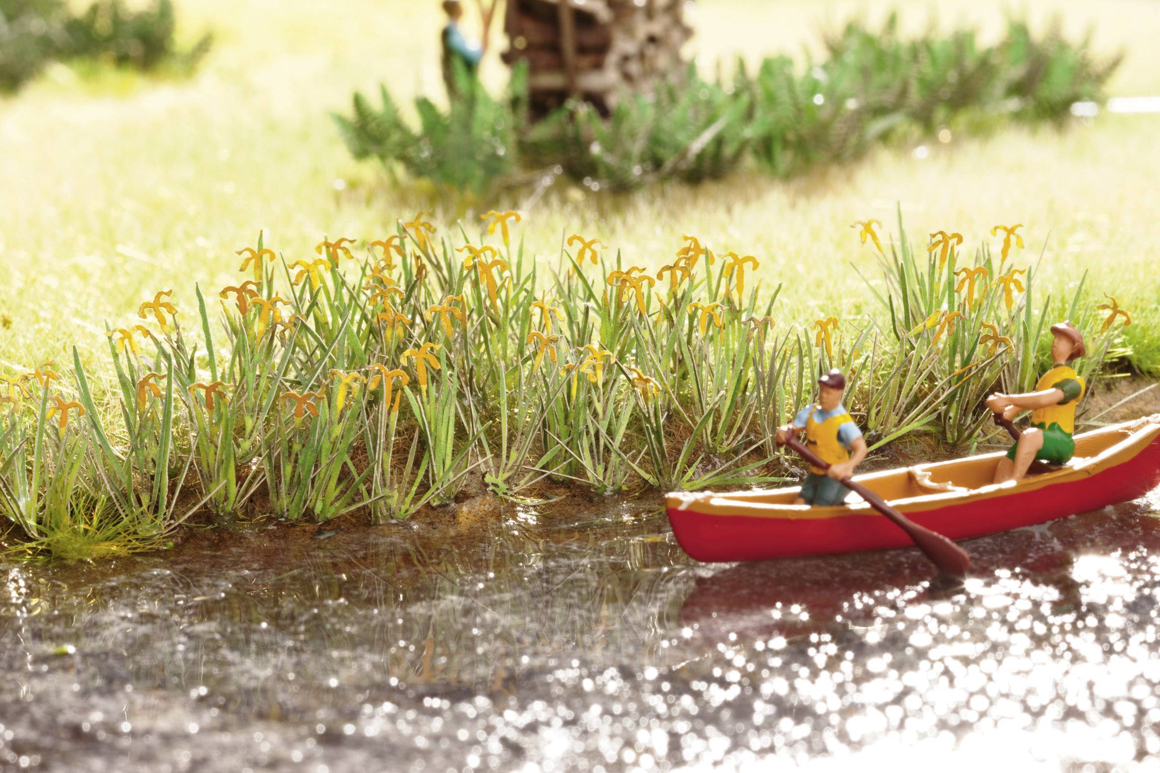 A red canoe with two people is paddling on a small river, surrounded by green banks with yellow flowers.
