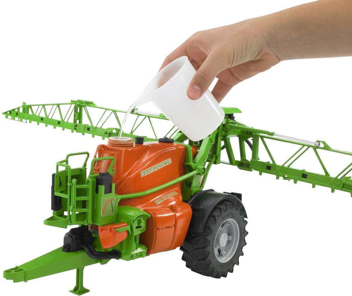 A hand is pouring liquid from a white cup into the tank of a green and orange agricultural model with tyres and arms.