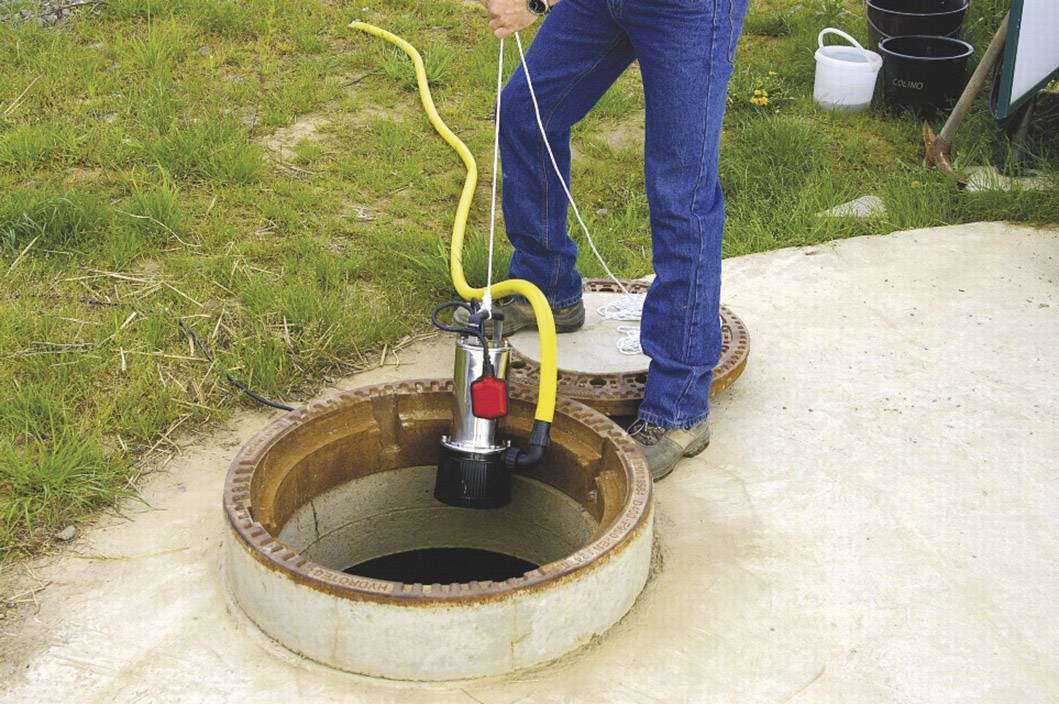A person is lowering a submersible pump into an open sewage pipe. The surrounding area is grassy, and various pieces of equipment are visible.