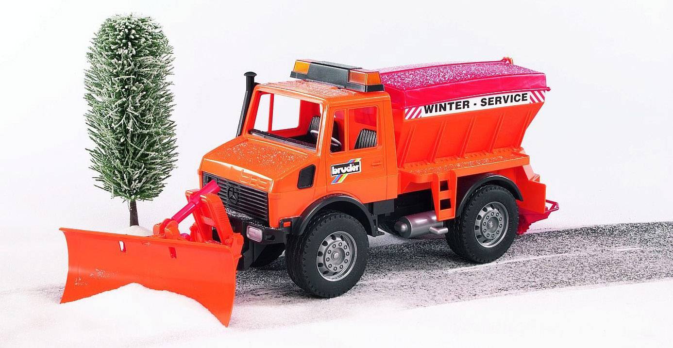 A red toy snow plough vehicle with the inscription 'Winter Service' drives on a snow-covered road beside a tree.