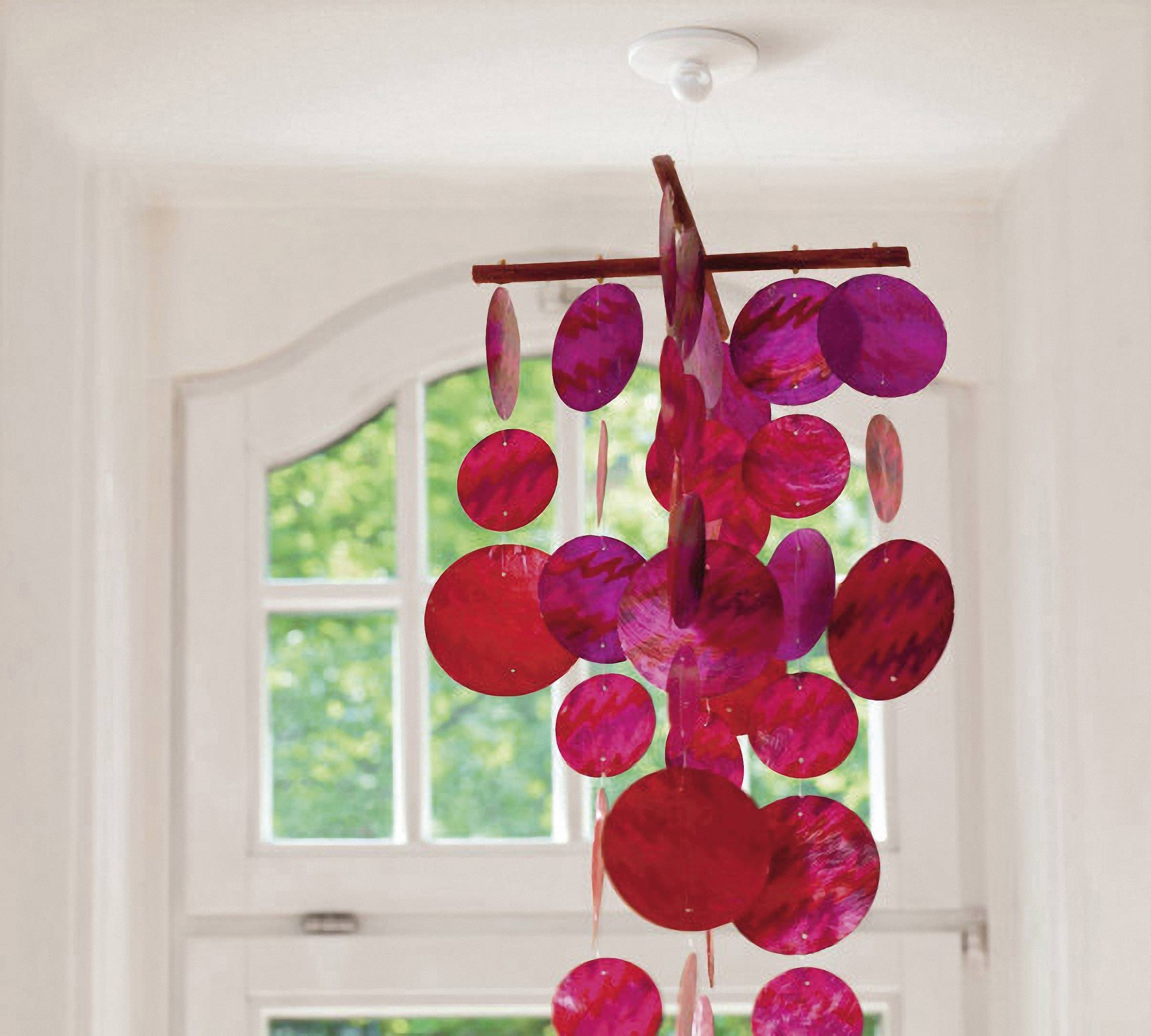 A mobile of red and purple discs hangs in front of a window with a green landscape in the background.