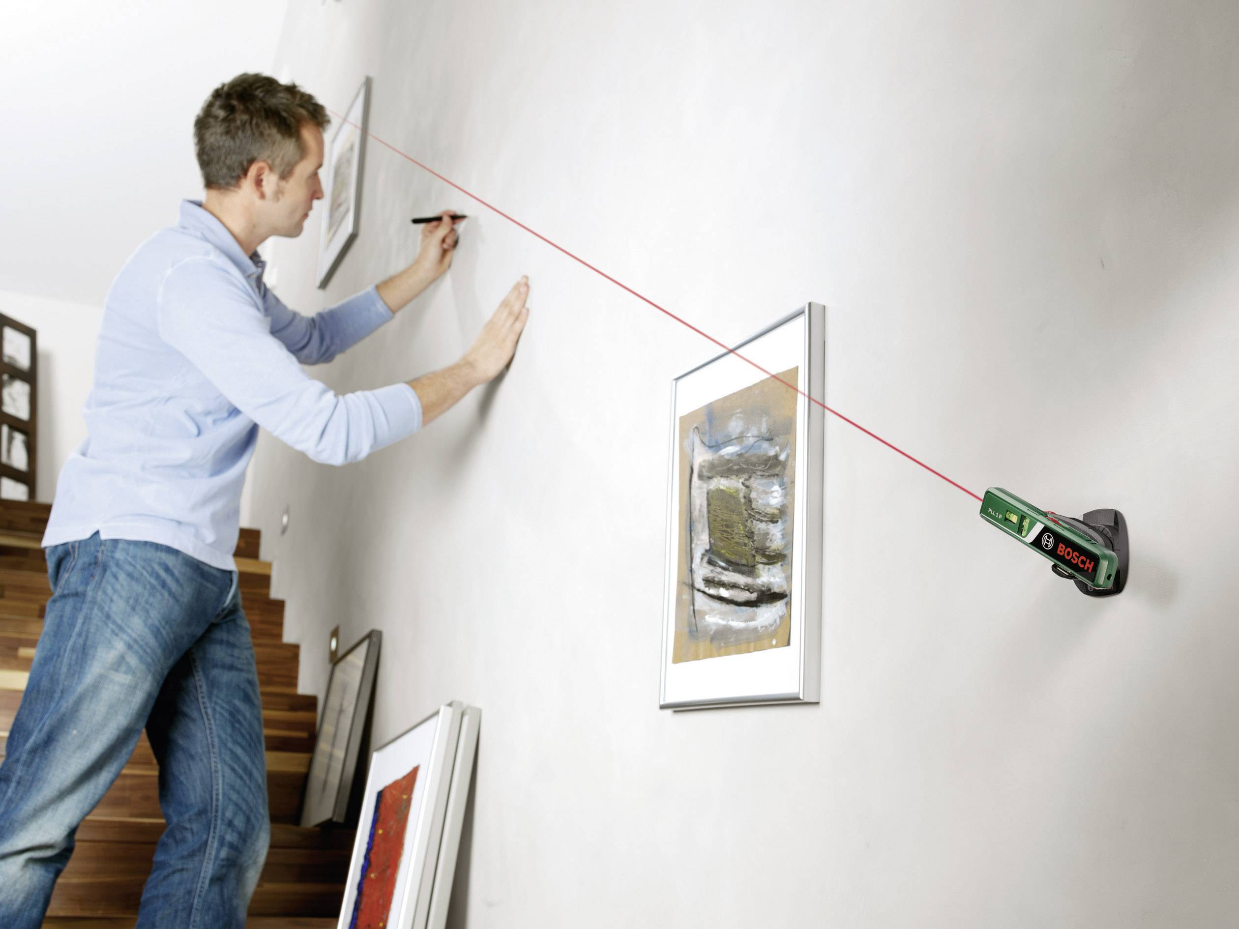 A man is mounting a picture on a wall using a laser tool attached to the wall to ensure straight alignment.