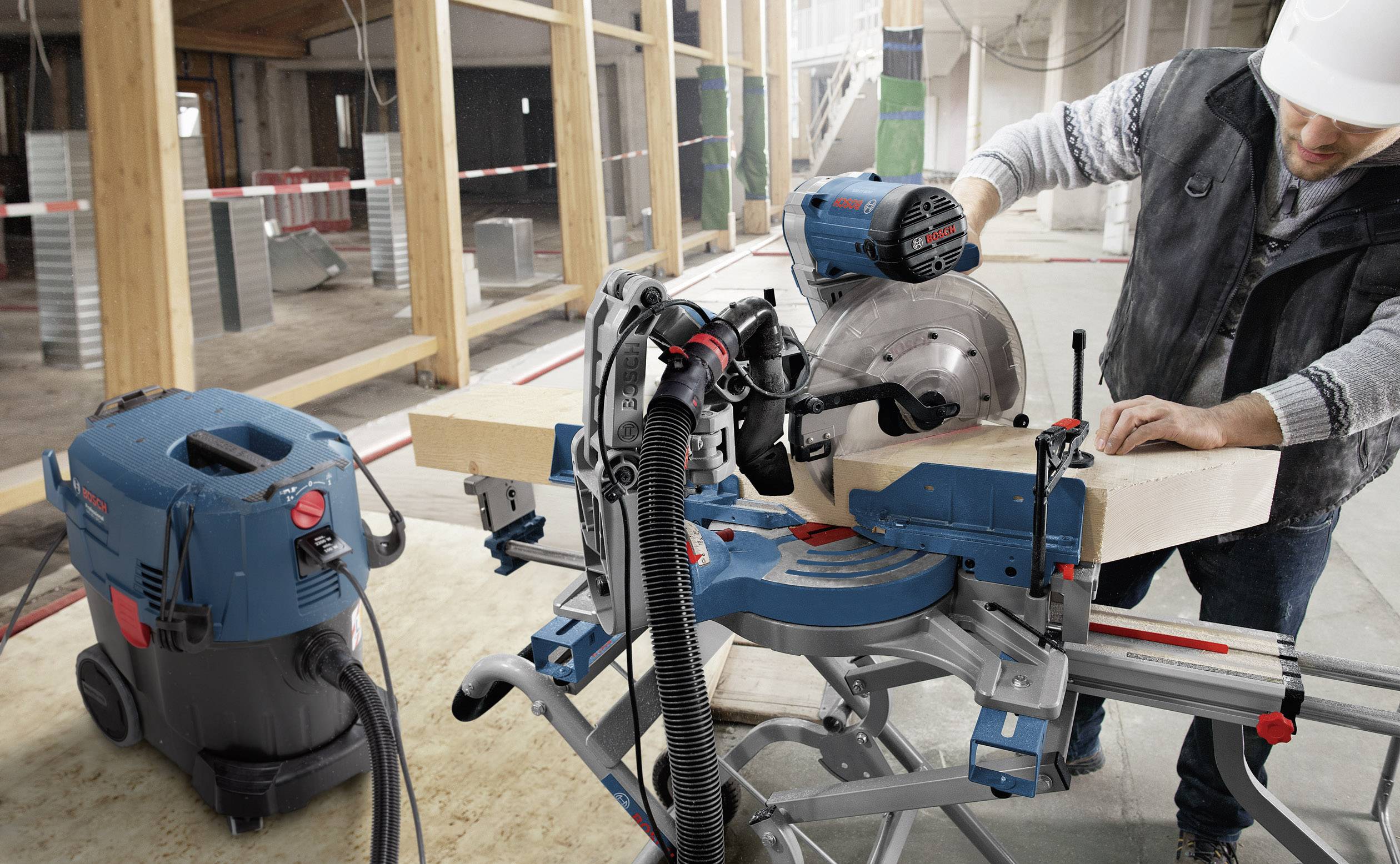 A worker is cutting a wooden board with a table circular saw in a workshop. An extraction system is connected to the saw.