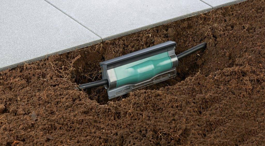 A green pipe set flush with the ground, with black connections on the left and right, beside a row of grey paving slabs.