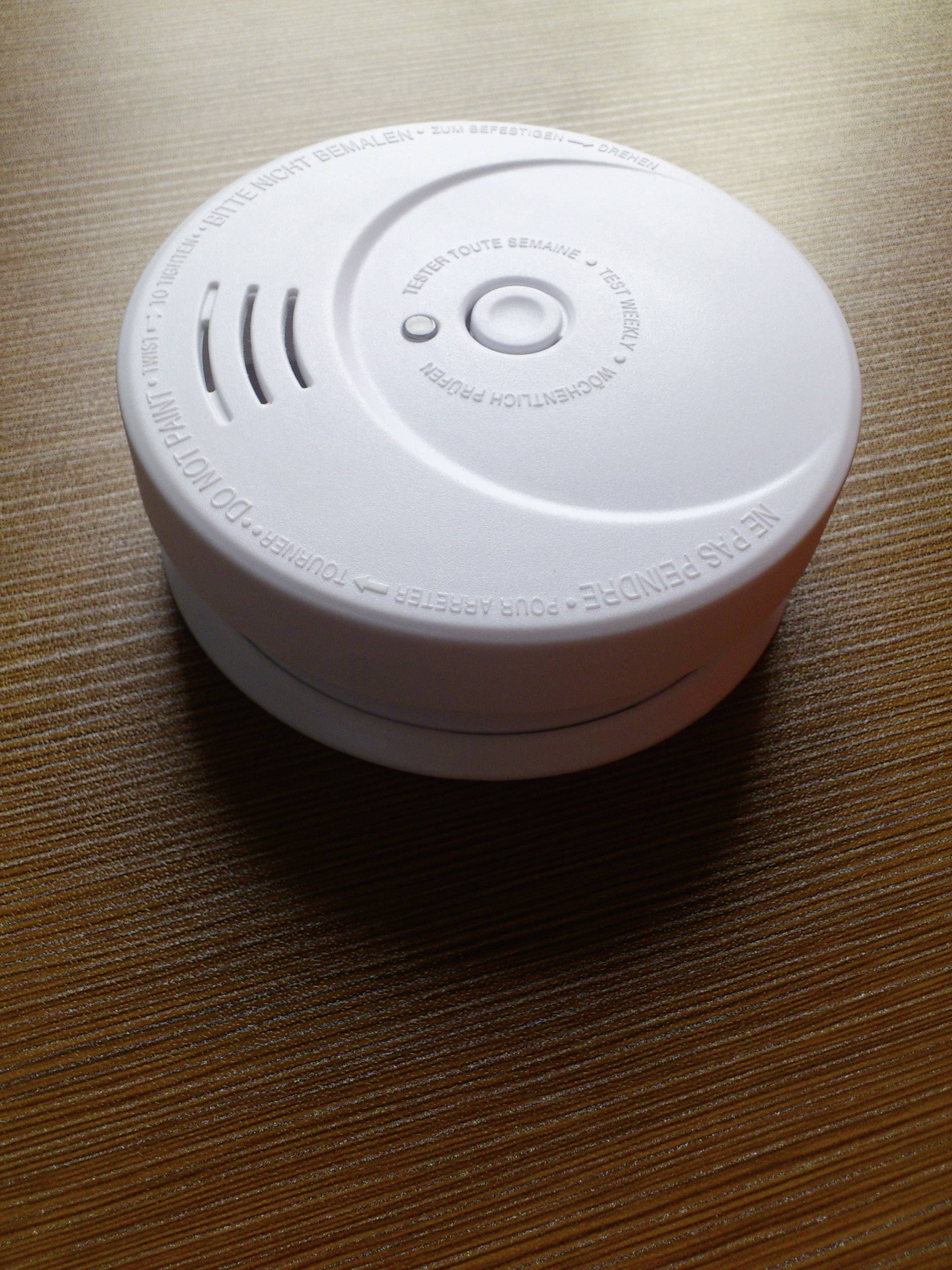 A smoke detector on a wooden table. The smoke detector has a smooth, white casing with a circular grille and a button.