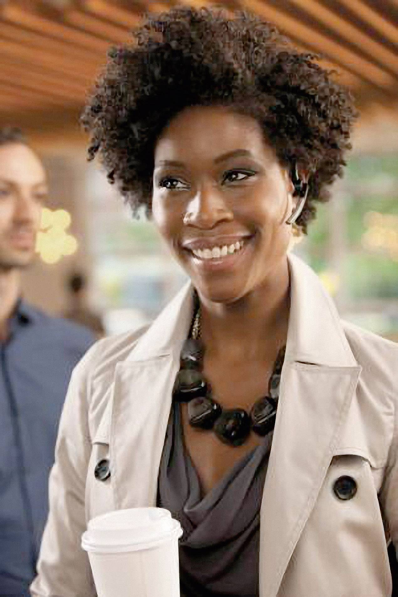 A smiling person with curly hair and a headset is holding a coffee mug. They are wearing a coat and a necklace. Blurred people are in the background.